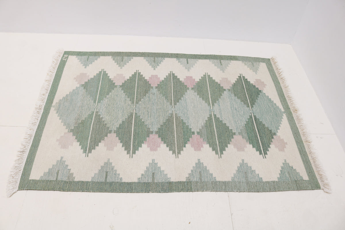 Angled view of Swedish Kilim rug featuring geometric diamond motifs in sage green and dusty pink