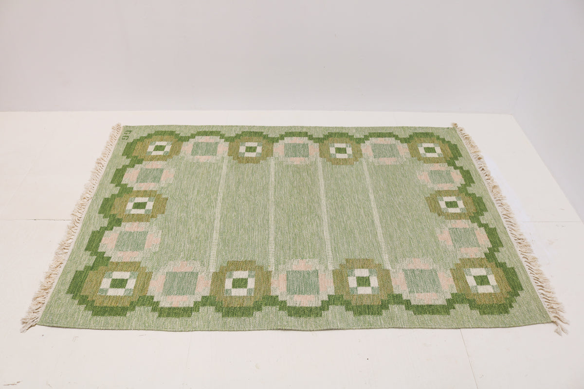 Angled view of Swedish Kilim rug featuring green, olive, ivory and pale pink geometric patterns