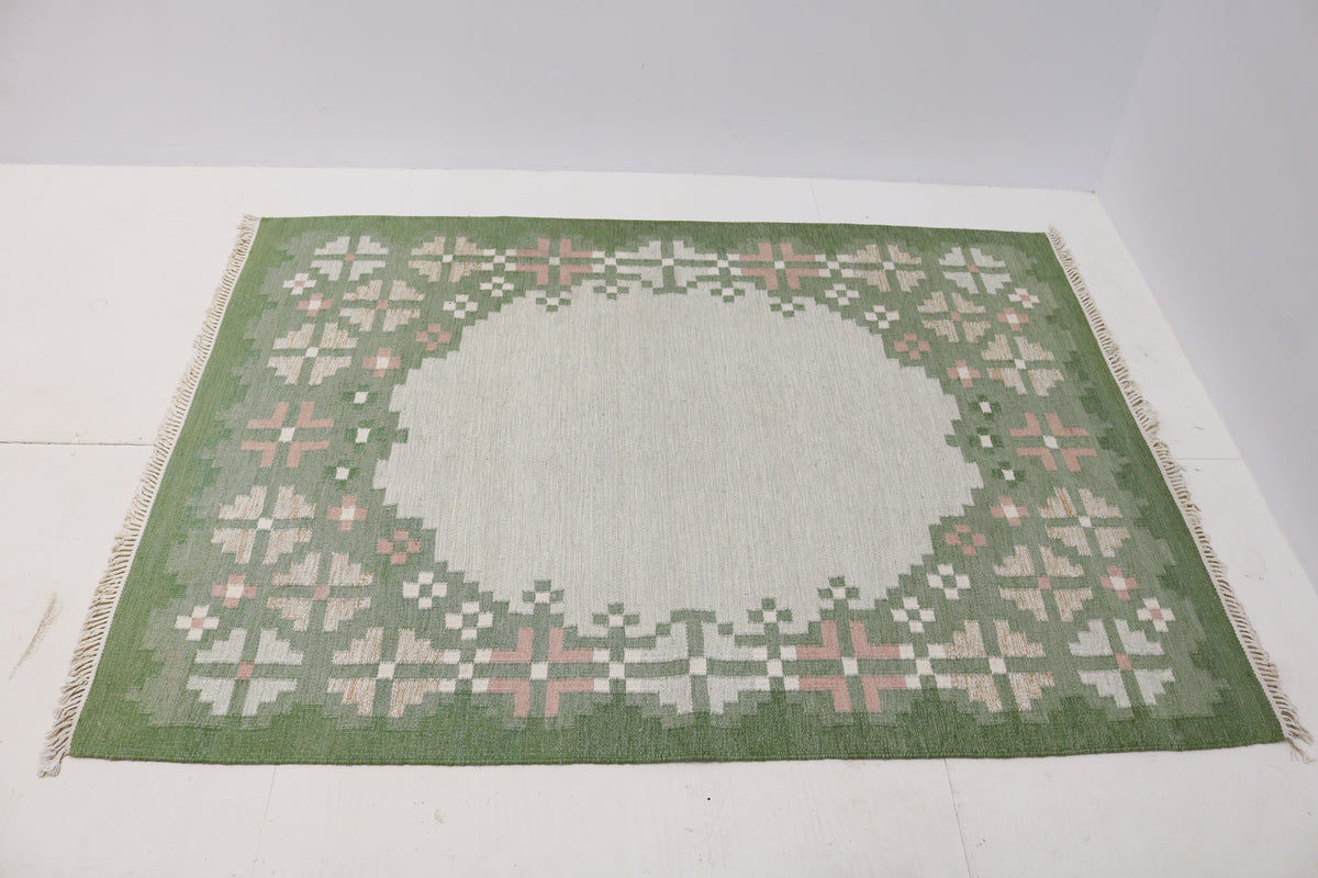 Angled view of Swedish Kilim rug featuring geometric floral motifs in sage green, ivory and pink