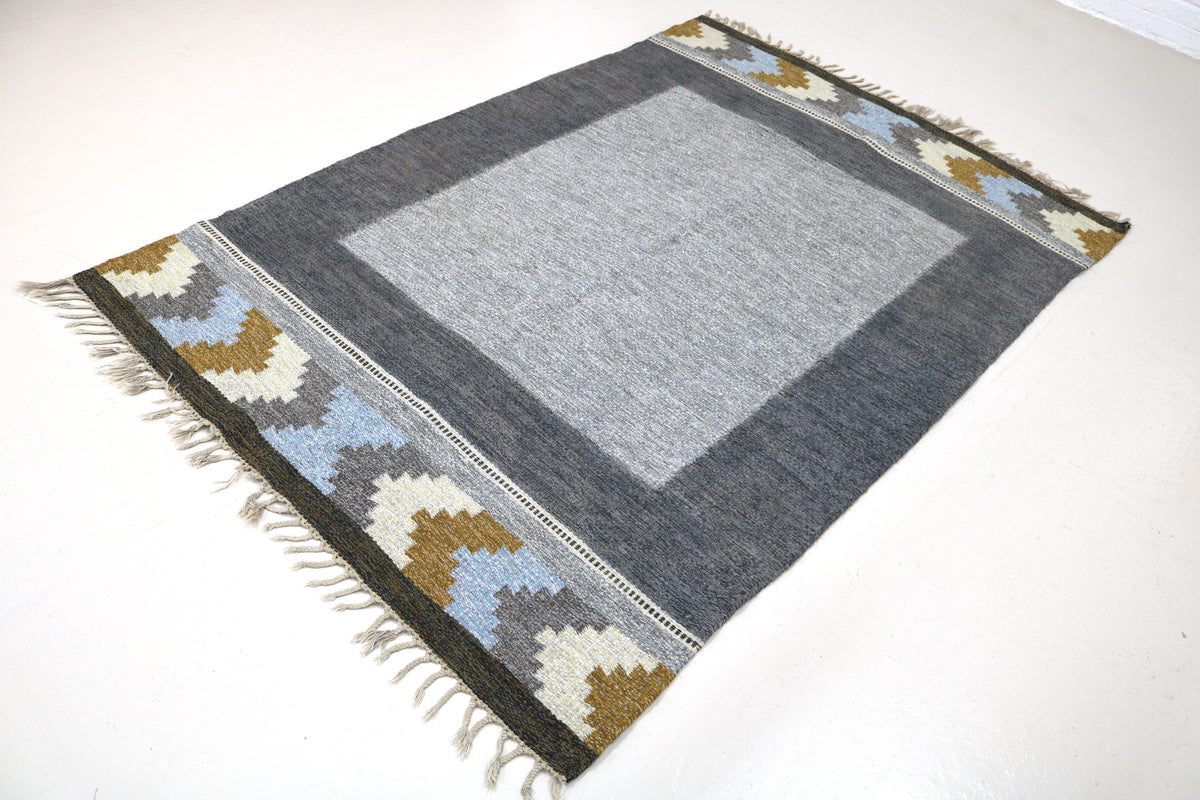 Angled view of Swedish Kilim rug featuring grey field and geometric border in muted colours
